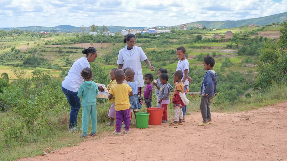 Children and adults gathered outdoors near buckets and a green landscape
